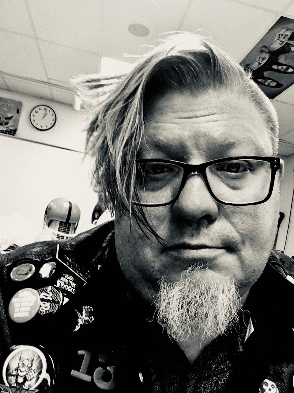 Portrait of Brent Elam. Image is black and white featuring Elam in his classroom. He is wearing a glasses and a jacket with pins on it.