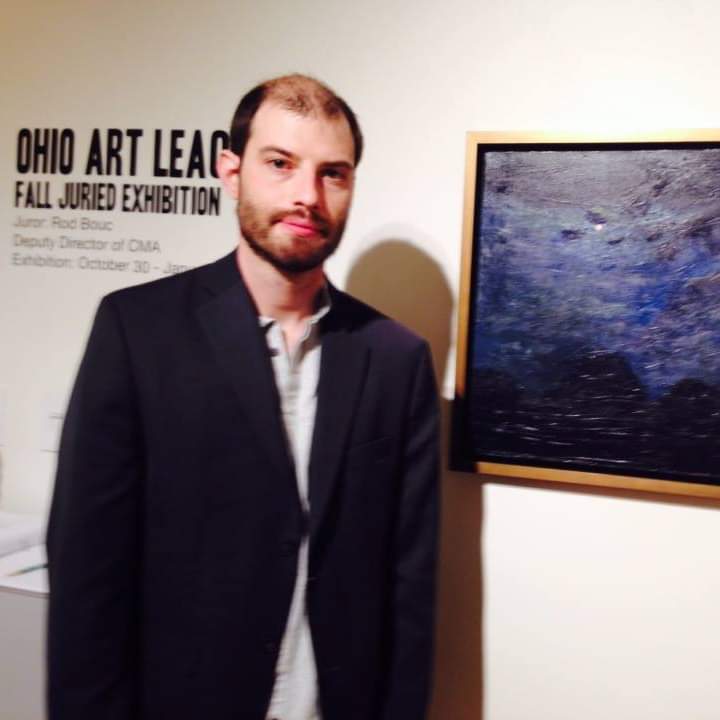 Portrait of Julian Cennamo next to a blue painting on a white wall.  Cennamo has a beard and is wearing a white shirt with a black jacket.