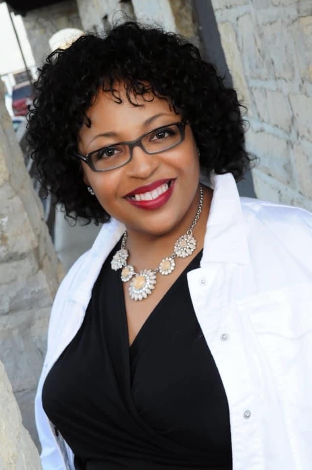 Portrait of Michol Childress. Michol has dark curly hair, is wearing glasses and a black shirt with a white jacket. The background is a stone building.